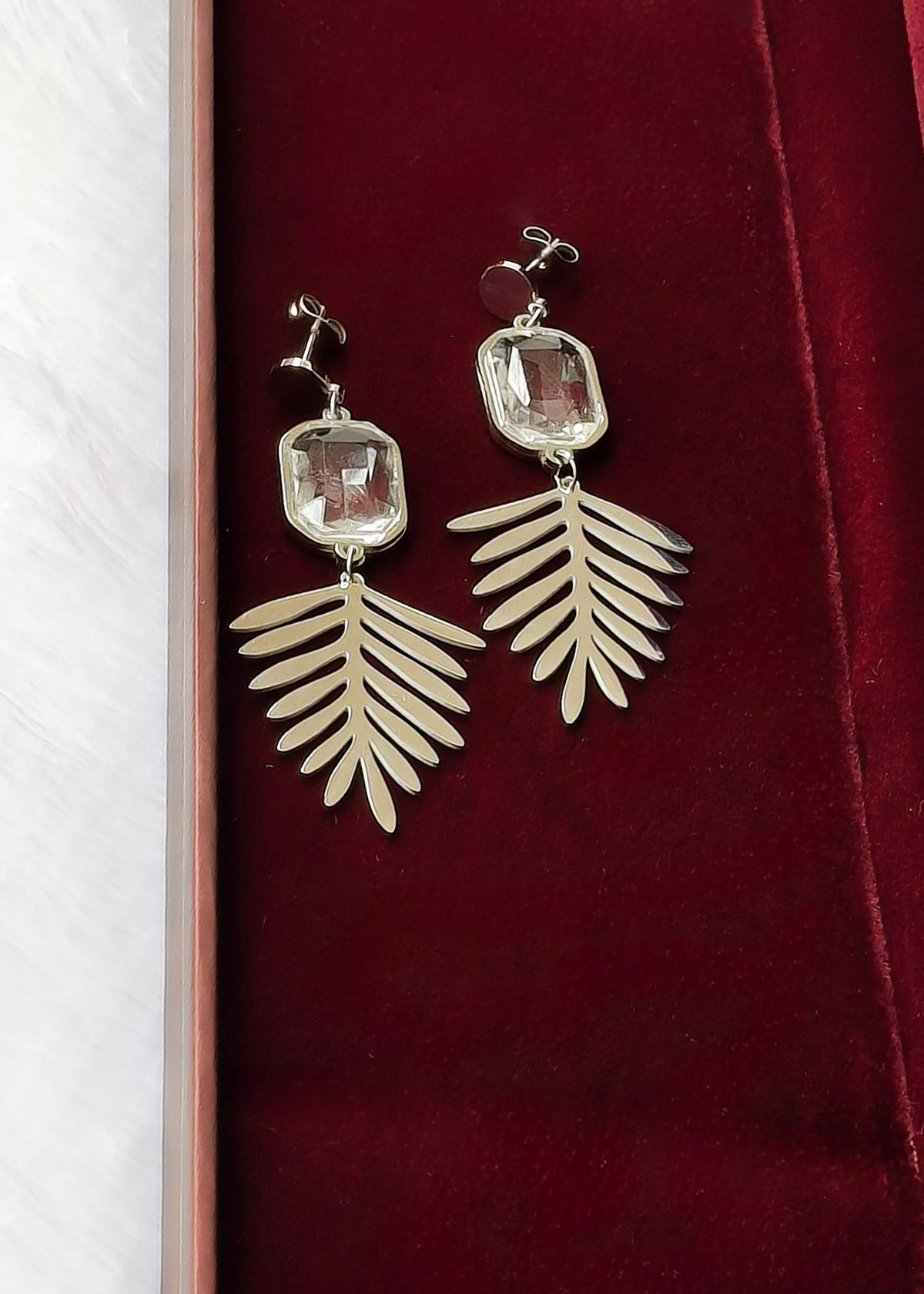 A pair of silver-toned drop earrings with rectangular clear gemstones and leaf-shaped dangling elements, displayed on a dark red velvet jewelry box.