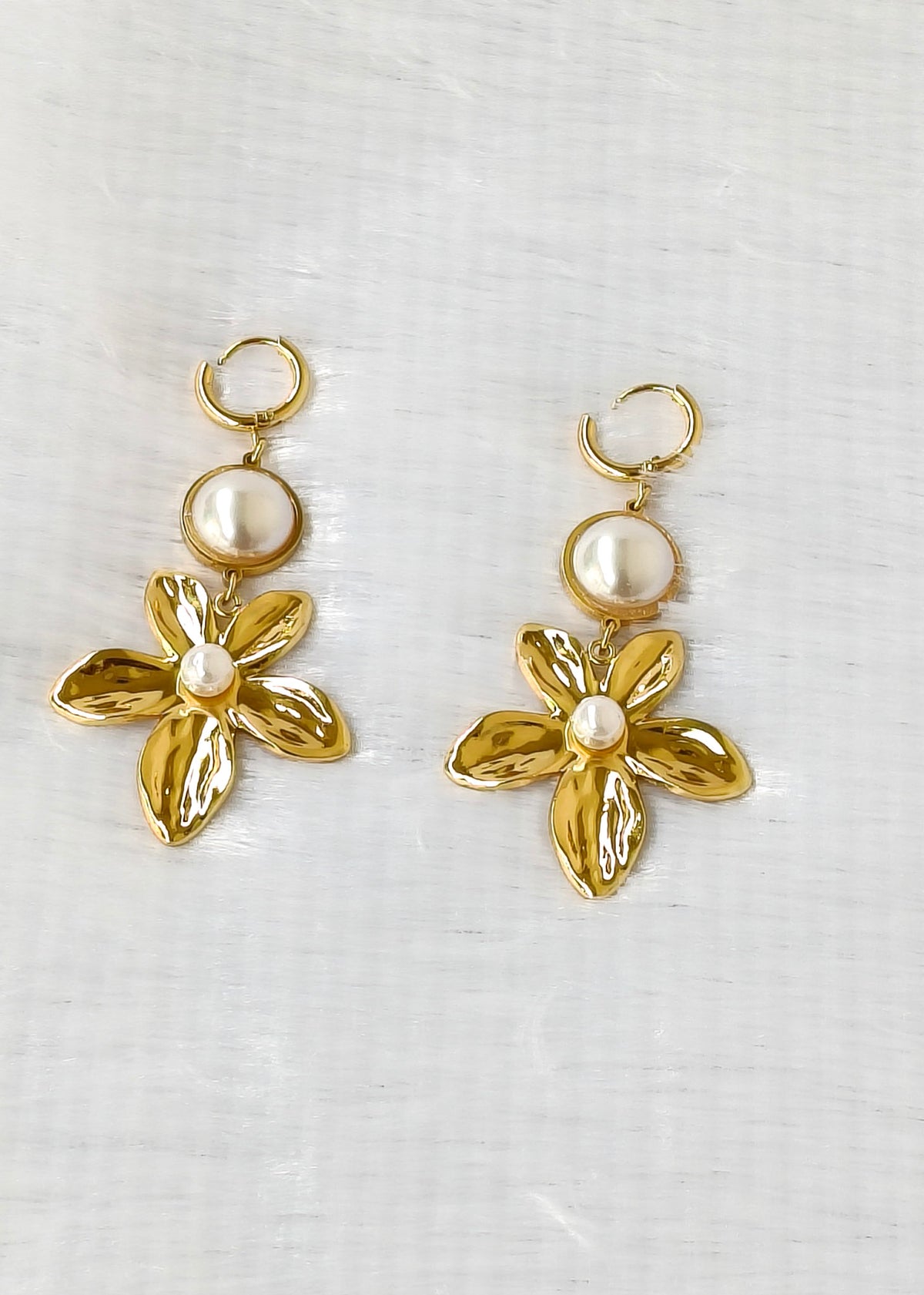 A pair of gold-tone dangle earrings featuring a small circular pearl-like bead above a five-petal flower with a small white pearl at the center, displayed on a light textured fabric background.