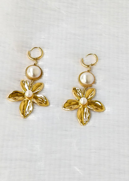 A pair of gold-tone dangle earrings featuring a small circular pearl-like bead above a five-petal flower with a small white pearl at the center, displayed on a light textured fabric background.