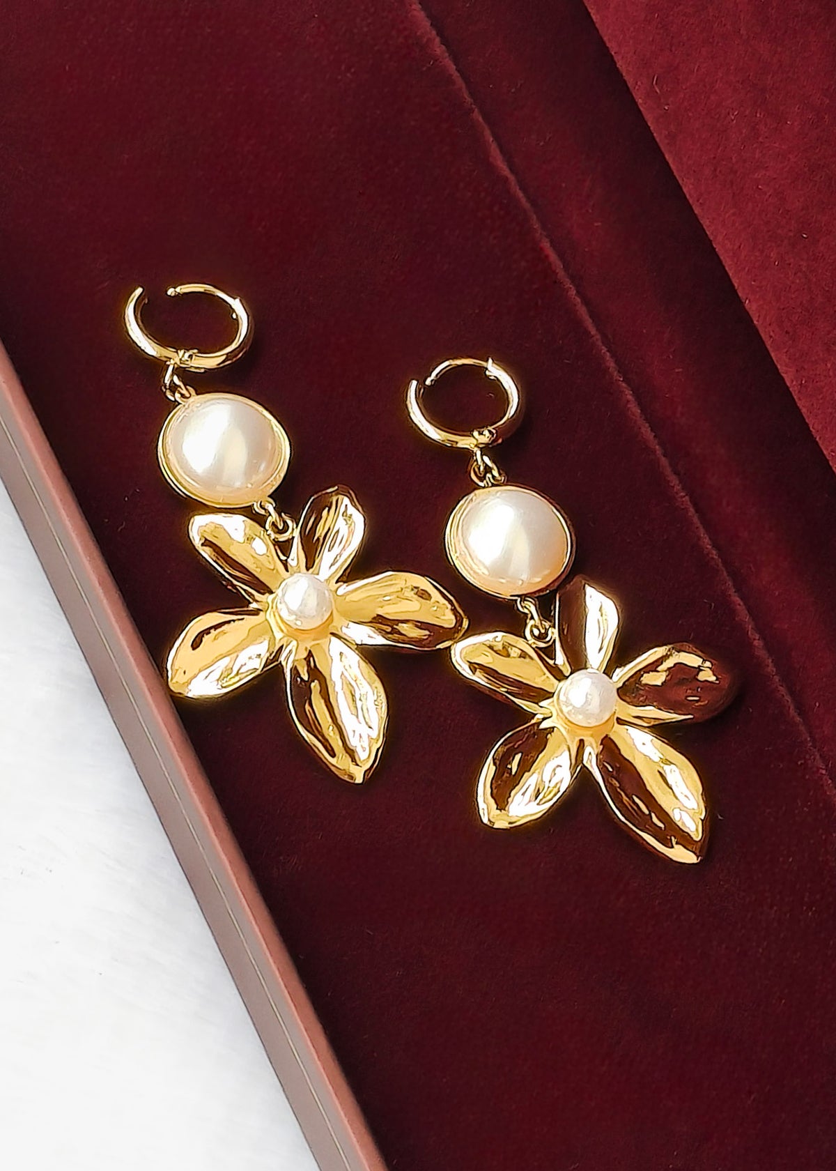 A pair of gold-tone flower drop earrings with round white faux-pearl beads, displayed on a deep burgundy velvet jewelry box.