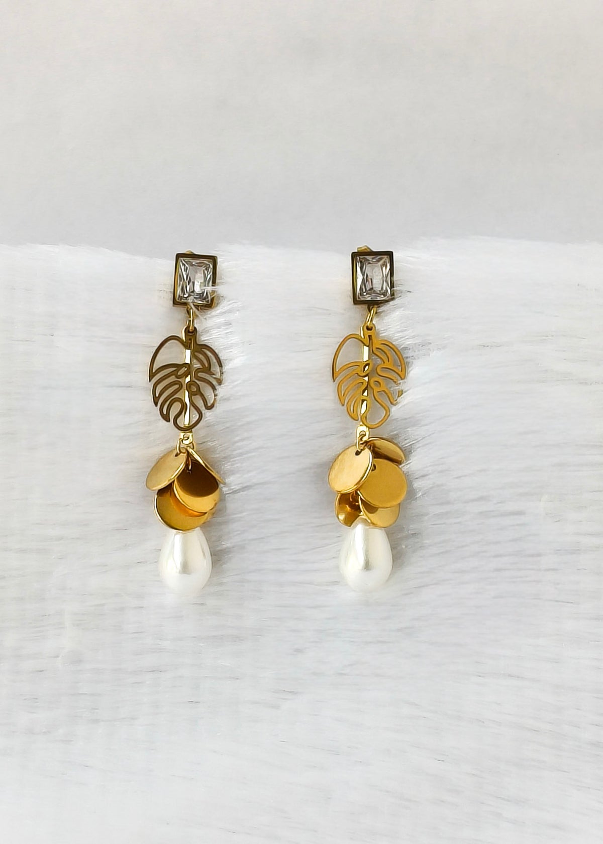 A pair of gold drop earrings with leaf filigree, gold disc charms, and white pearl-like drops, displayed on a white textured surface.