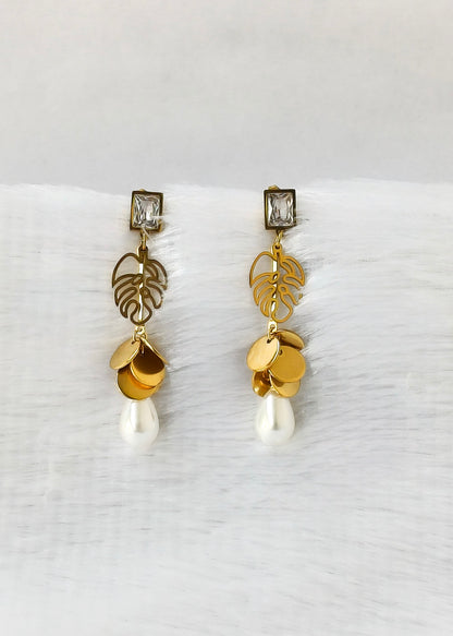A pair of gold drop earrings with leaf filigree, gold disc charms, and white pearl-like drops, displayed on a white textured surface.