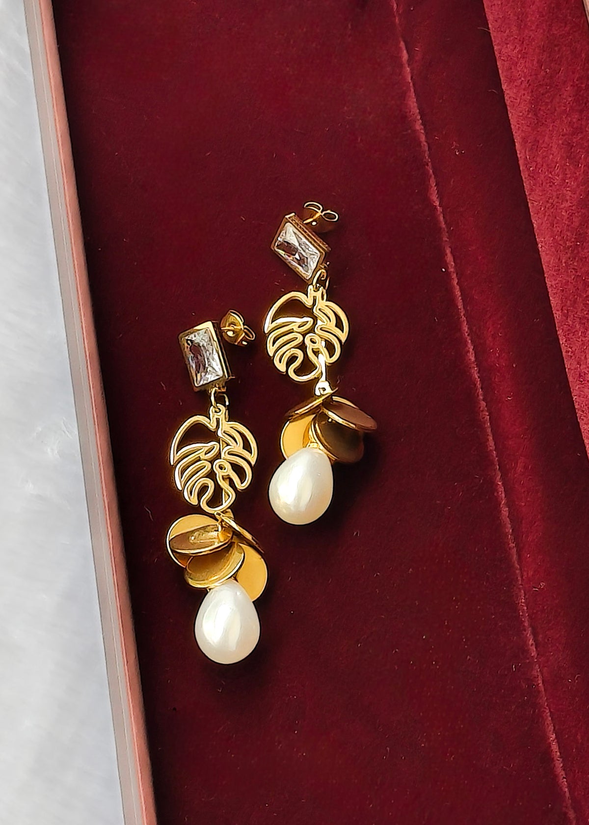 A pair of gold and white drop earrings displayed on a deep red velvet jewelry box. The earrings feature a gold leaf design, gold coin charms, and white teardrop pearls.