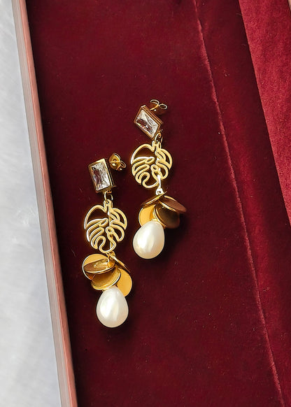 A pair of gold and white drop earrings displayed on a deep red velvet jewelry box. The earrings feature a gold leaf design, gold coin charms, and white teardrop pearls.