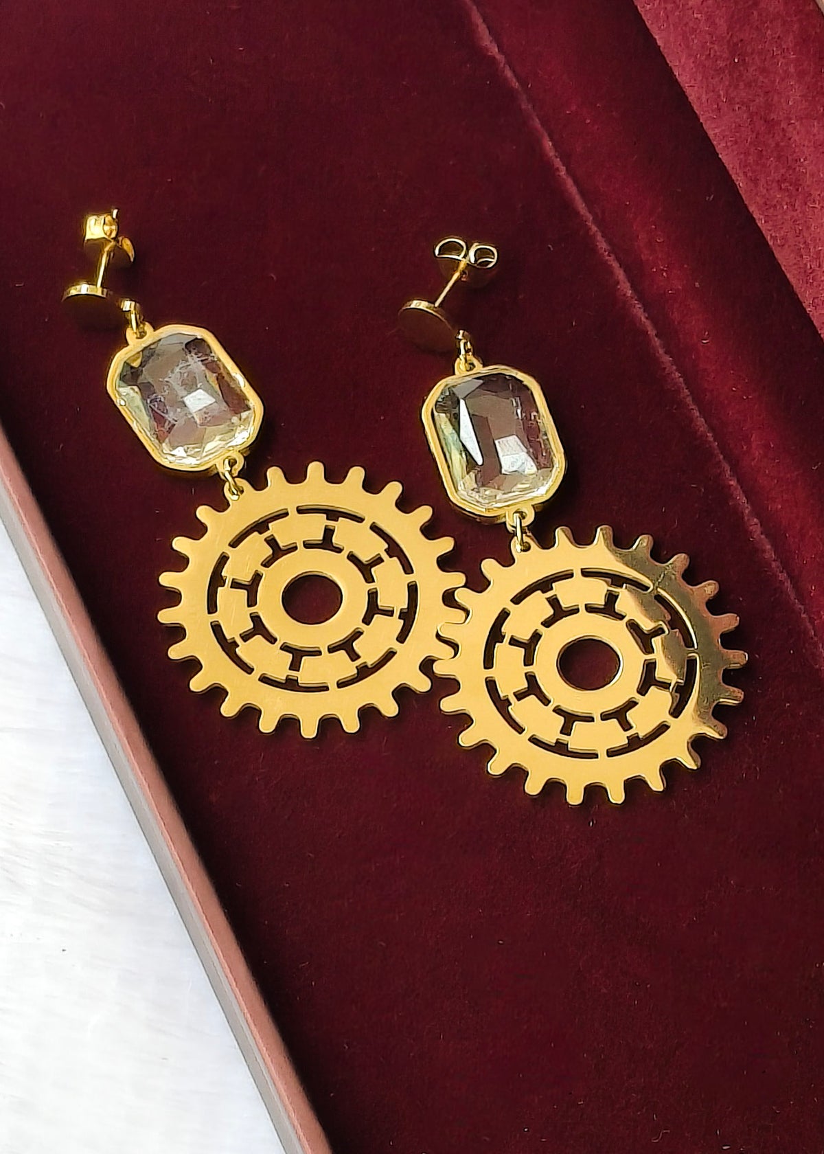 A pair of gold-toned dangle earrings with rectangular clear gemstones and large gear-shaped cutout pendants, displayed on a deep red velvet jewelry box.