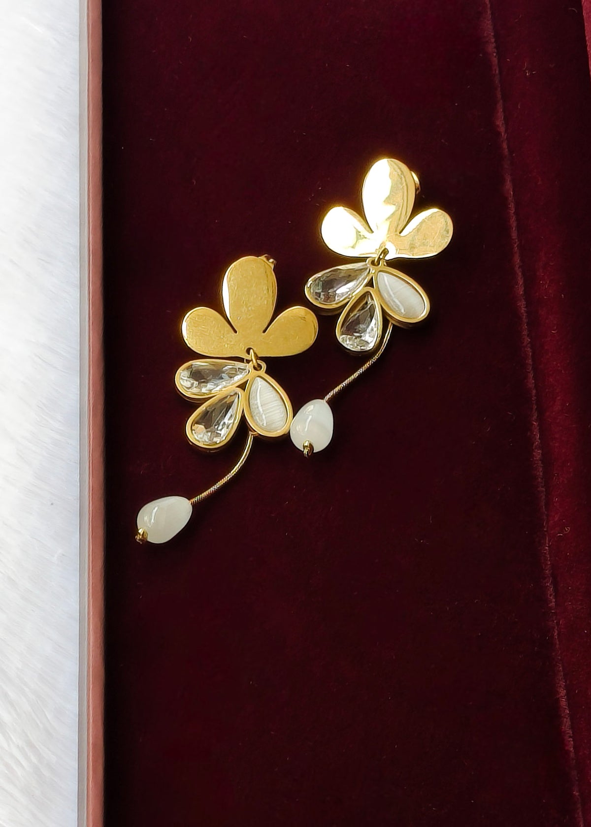 A pair of gold-tone flower-shaped earrings with clear faceted teardrop rhinestones and small white bead drops, displayed on a dark red velvet surface.