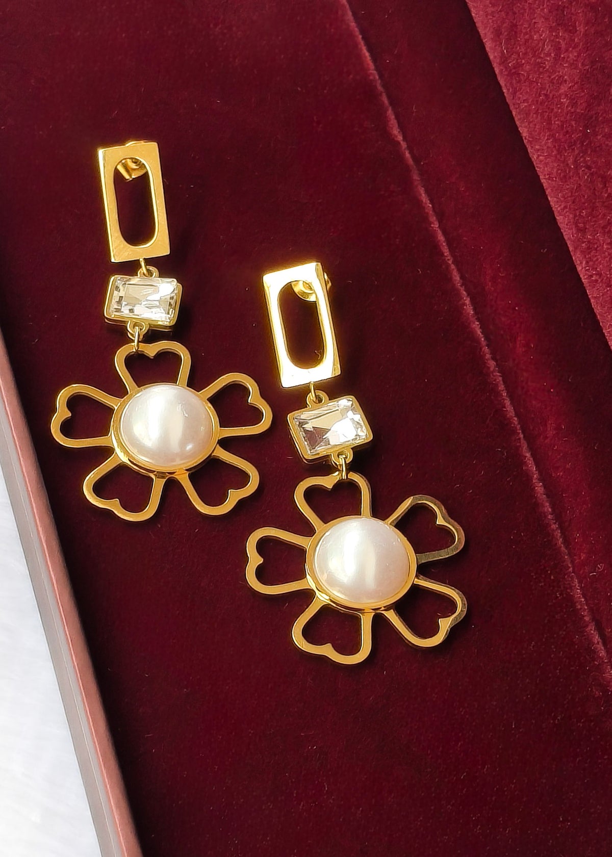 A pair of gold daisy drop earrings with white faux pearls in the center and clear rectangular crystals above, displayed on a deep red velvet jewelry box.