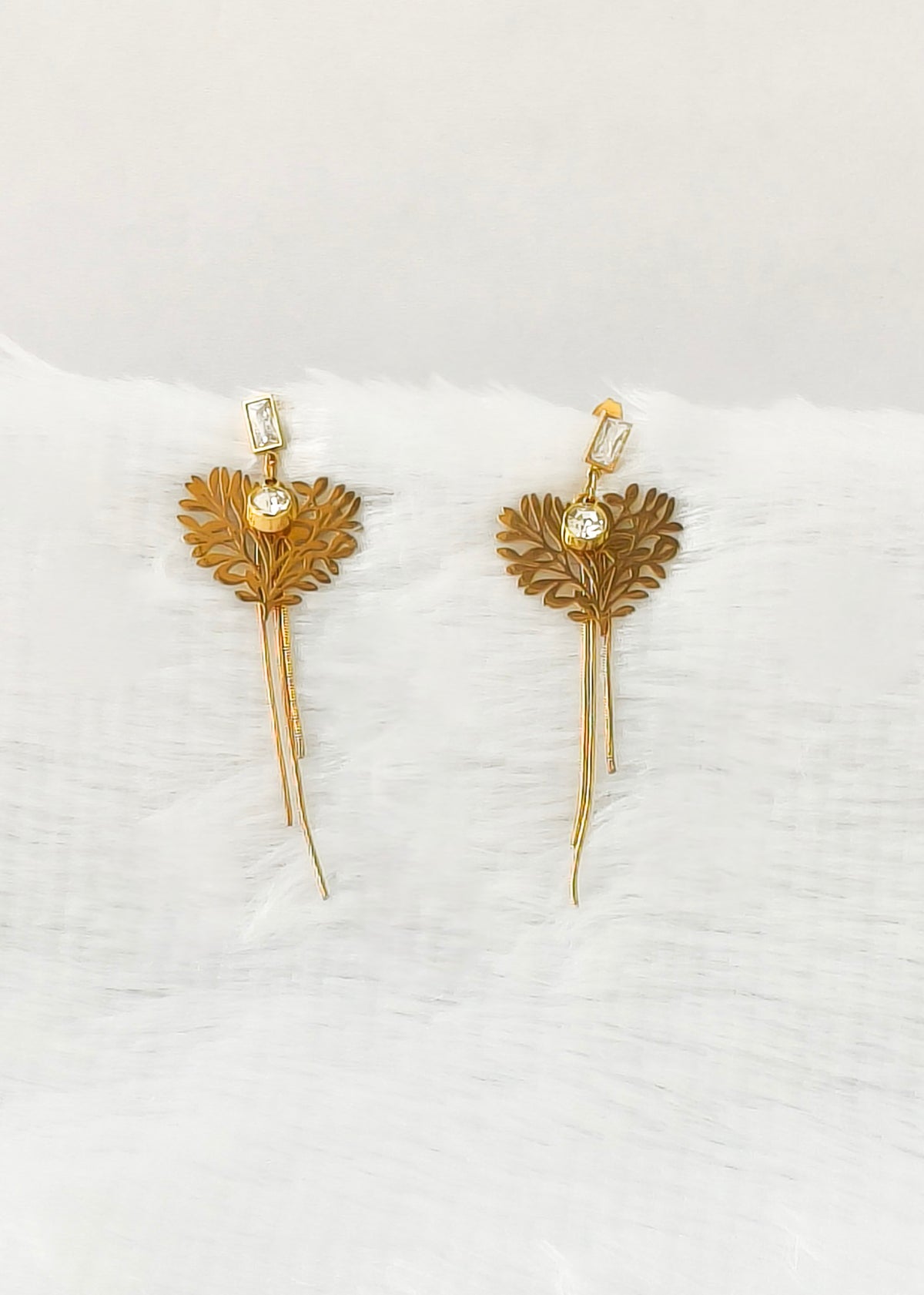 A pair of gold leaf drop earrings with small clear rectangular crystals and round clear center stones, displayed on a soft white textured surface.