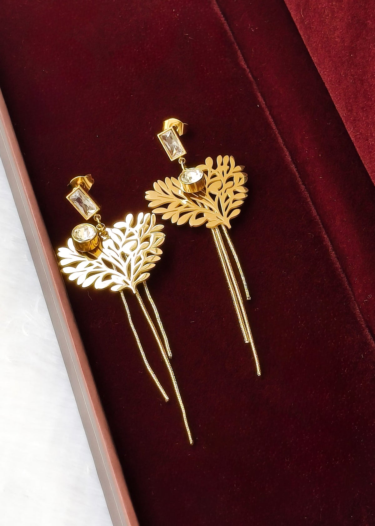 A pair of gold dangle earrings with leaf-shaped filigree pendants, rectangular clear gemstones, and long dangling chains, displayed on a deep red velvet jewelry box.