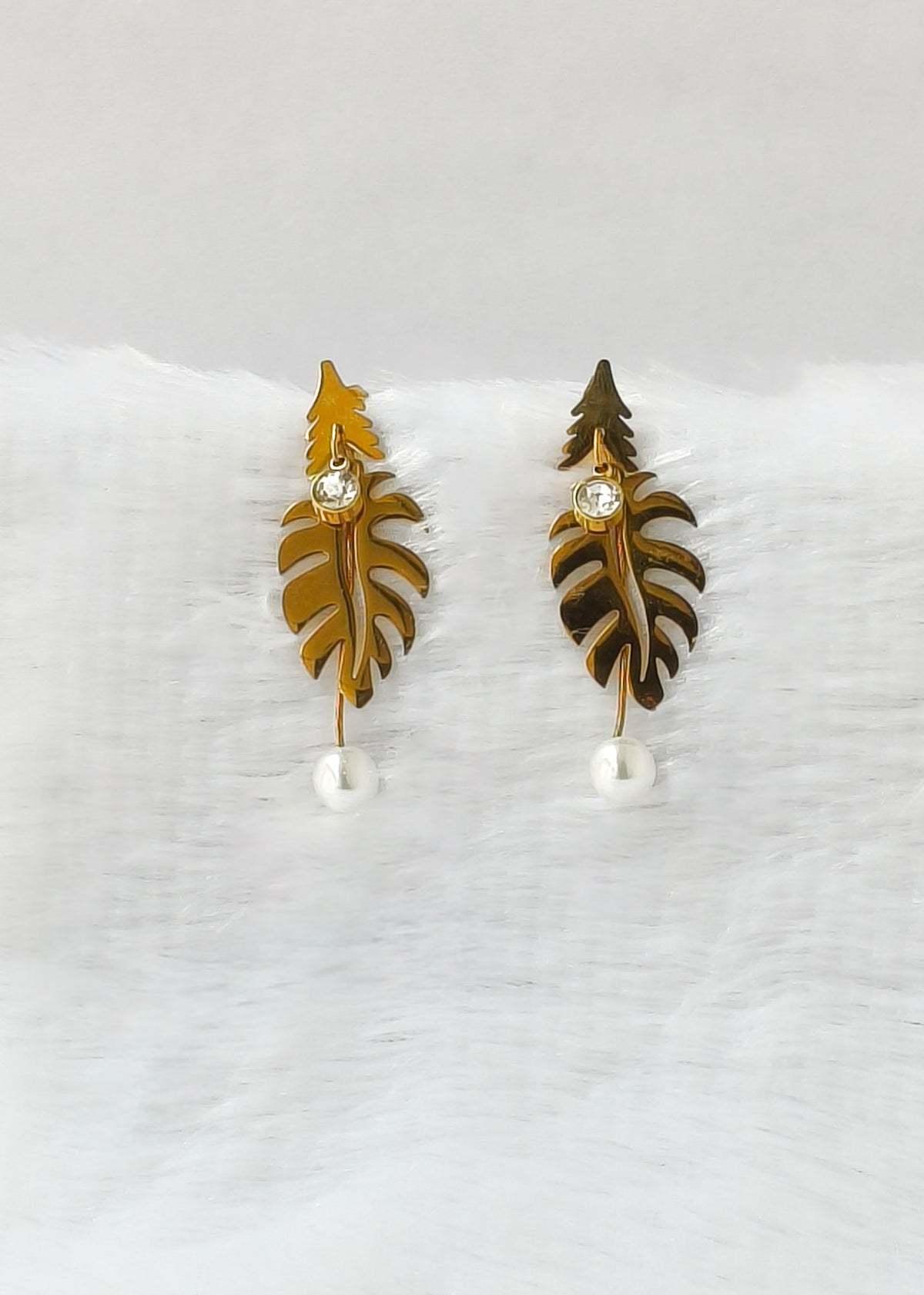 A pair of gold-toned leaf-shaped drop earrings with a small clear rhinestone in the center and a white pearl dangling at the bottom, displayed on a soft white textured surface.