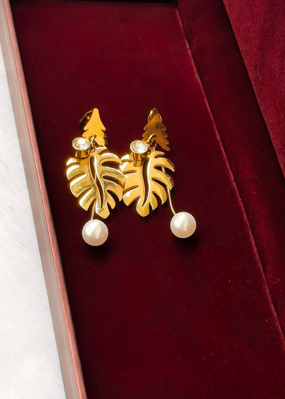 A pair of gold leaf drop earrings with white pearl beads hanging from them, displayed inside a dark red velvet jewelry box.
