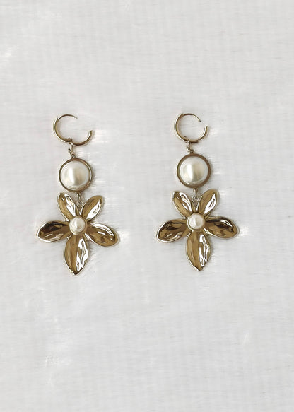 A pair of gold-tone drop earrings featuring a circular pearl-like bead and a textured flower charm with a small white pearl center, displayed on a light fabric background.