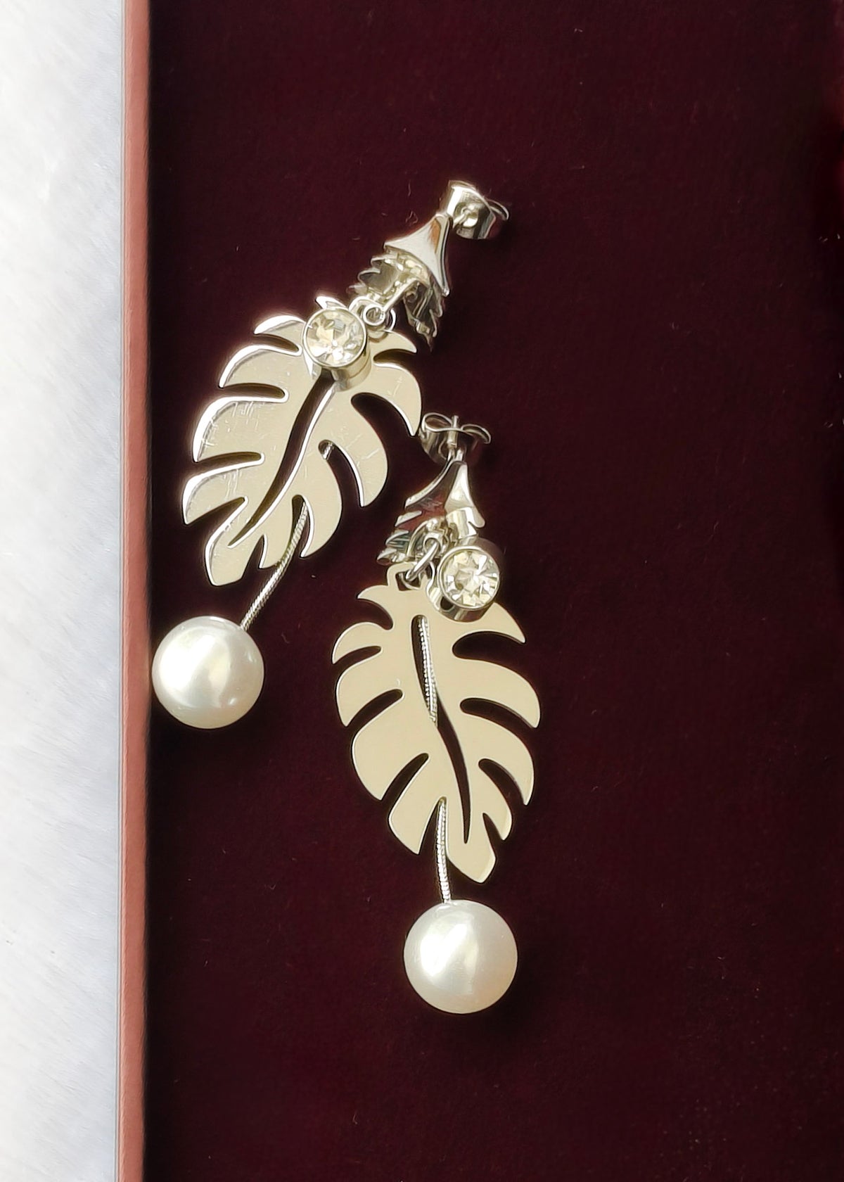 A pair of silver-toned dangle earrings with cutout leaf shapes, clear rhinestone accents, and white pearl drops, displayed on a dark burgundy velvet surface.