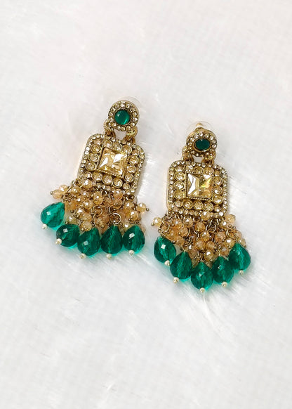 A pair of gold-toned chandelier earrings embellished with clear stones and hanging emerald green teardrop beads, displayed on a white textured background.
