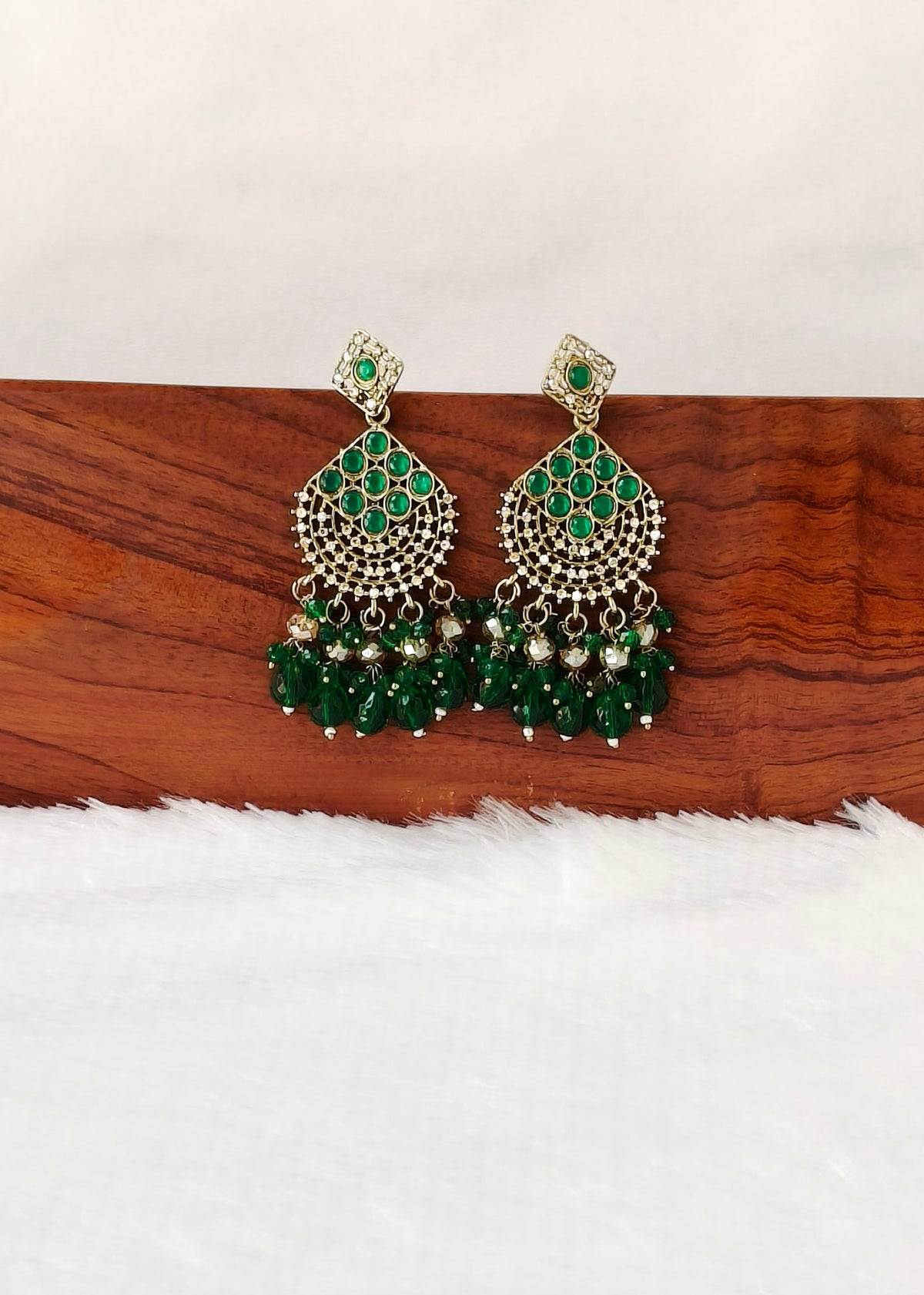 A pair of gold-plated Indian jhumka earrings with green stone embellishments and dangling green bead chains, displayed on a wooden block over a white faux fur surface.