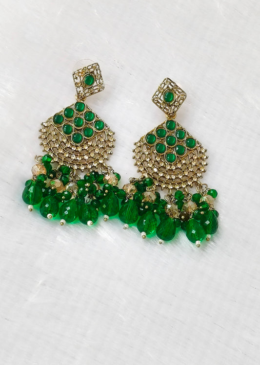 A pair of gold-plated chandelier earrings embellished with green stones and hanging green bead drops, displayed on a textured white background.