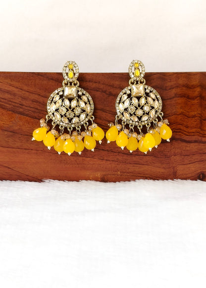 A pair of ornate gold-toned Indian jhumka earrings with yellow bead drops and clear rhinestone embellishments, displayed on a wooden block over a white textured surface.