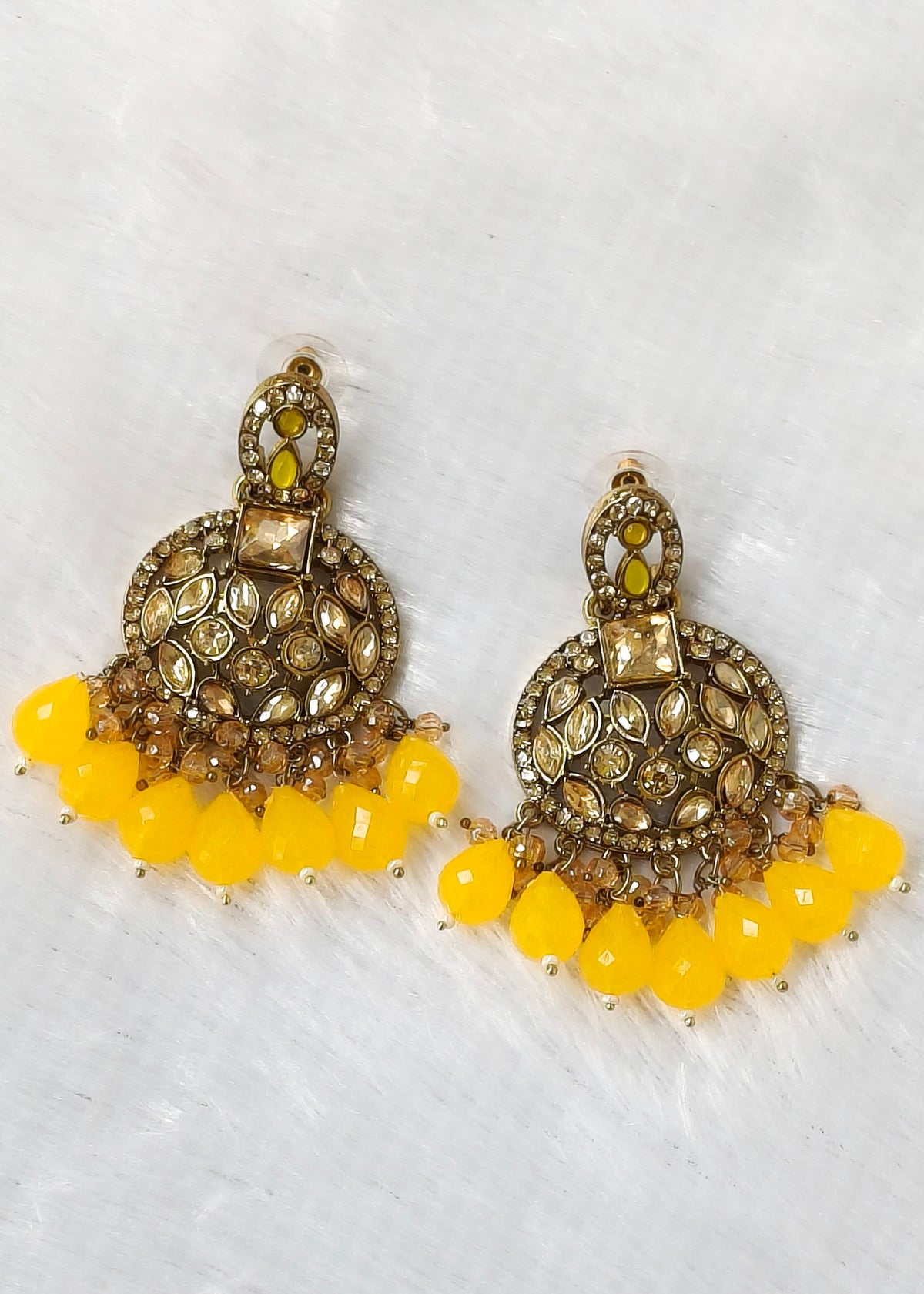 A pair of ornate gold-toned jhumka earrings with yellow teardrop beads and small yellow-green stones, displayed on a white textured background.