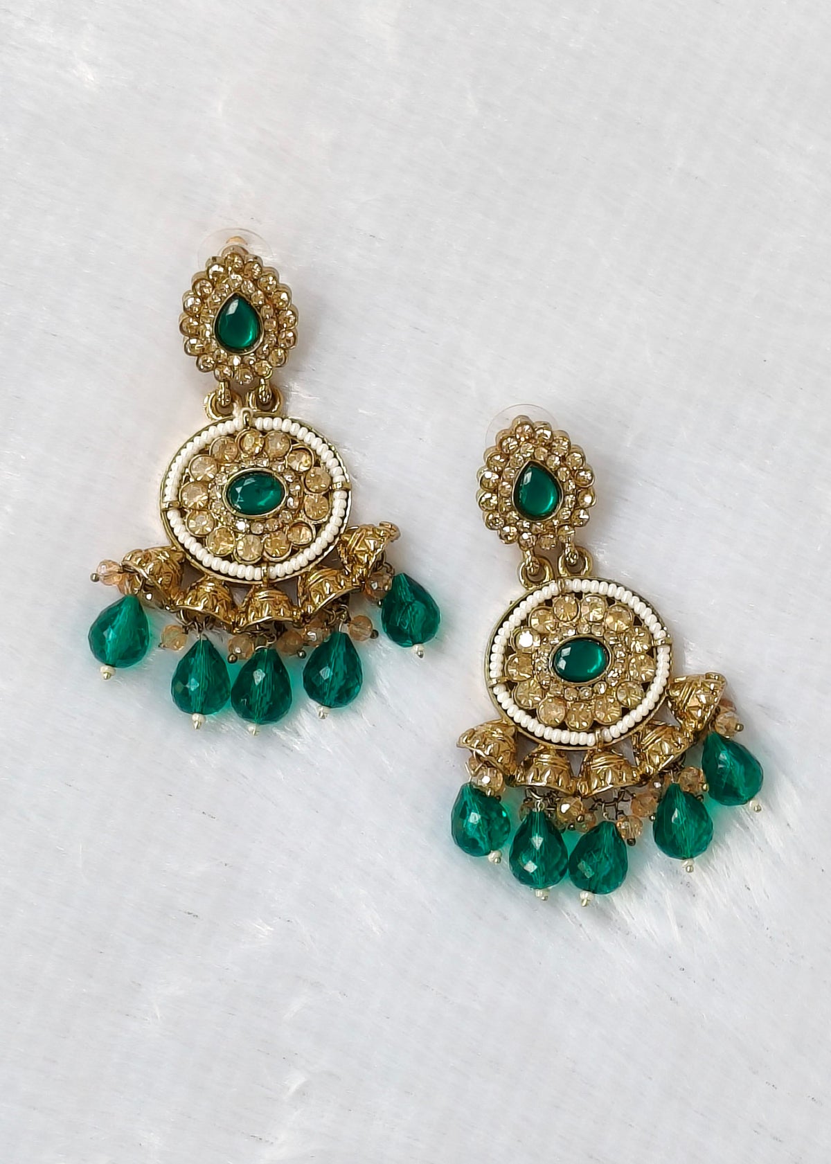 A pair of ornate gold-toned chandelier earrings with green gemstone accents and dangling teardrop beads, displayed on a textured white surface.