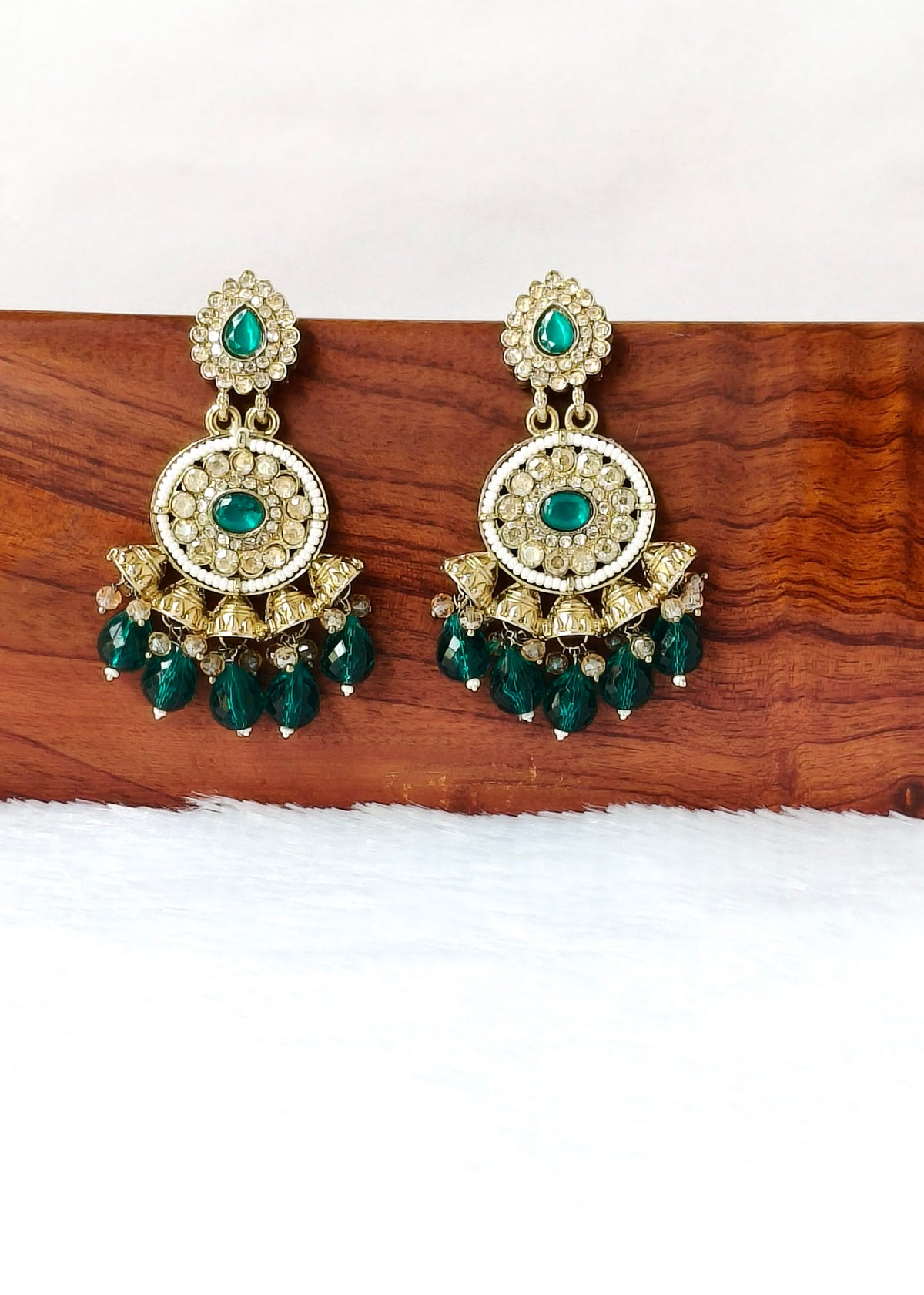 A pair of ornate gold-toned chandelier earrings with green stones and crystal-like accents, displayed on a wooden block against a white background.