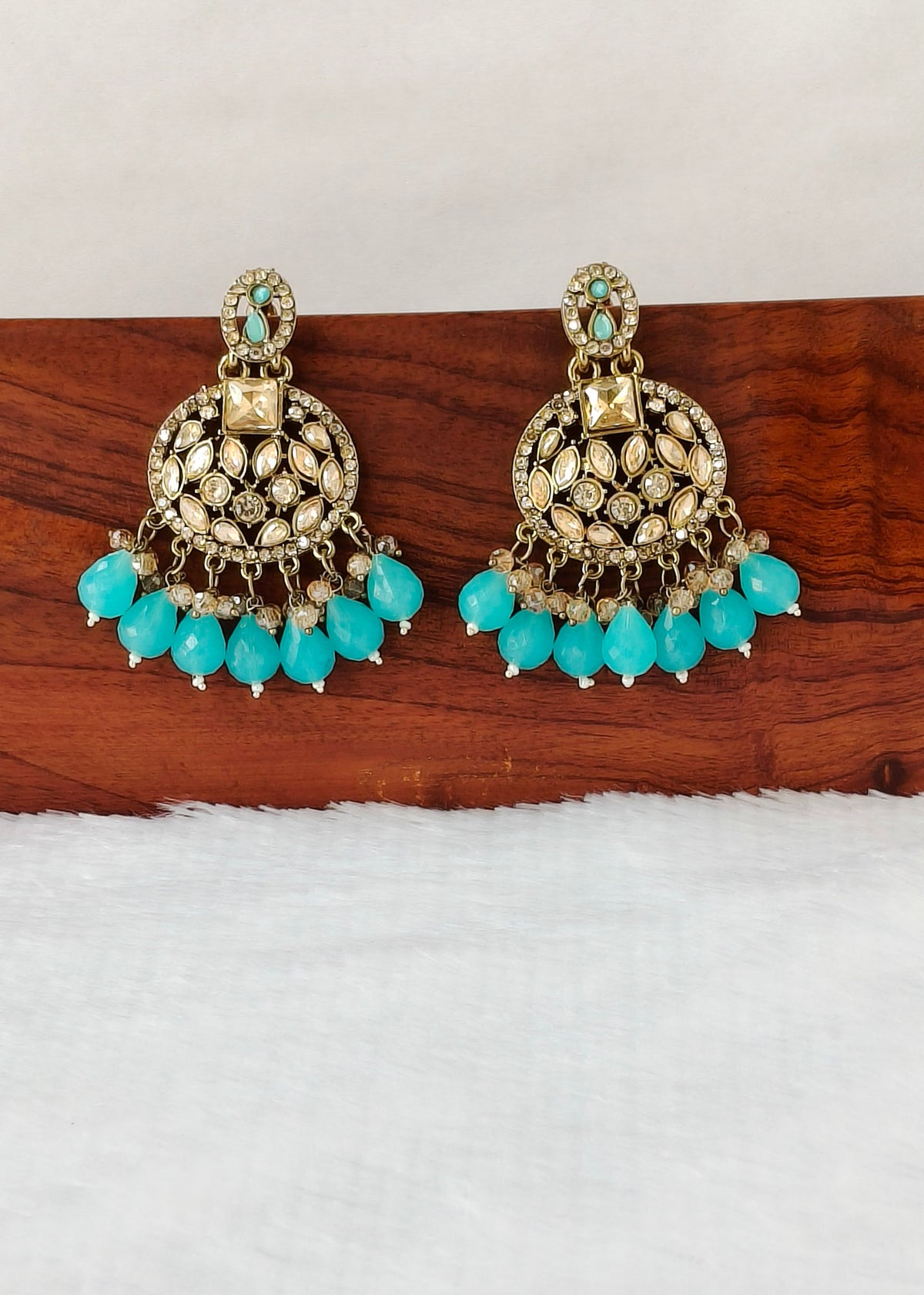 A pair of ornate gold-toned chandelier earrings with turquoise blue bead drops and small clear stones, displayed on a wooden block against a white background.