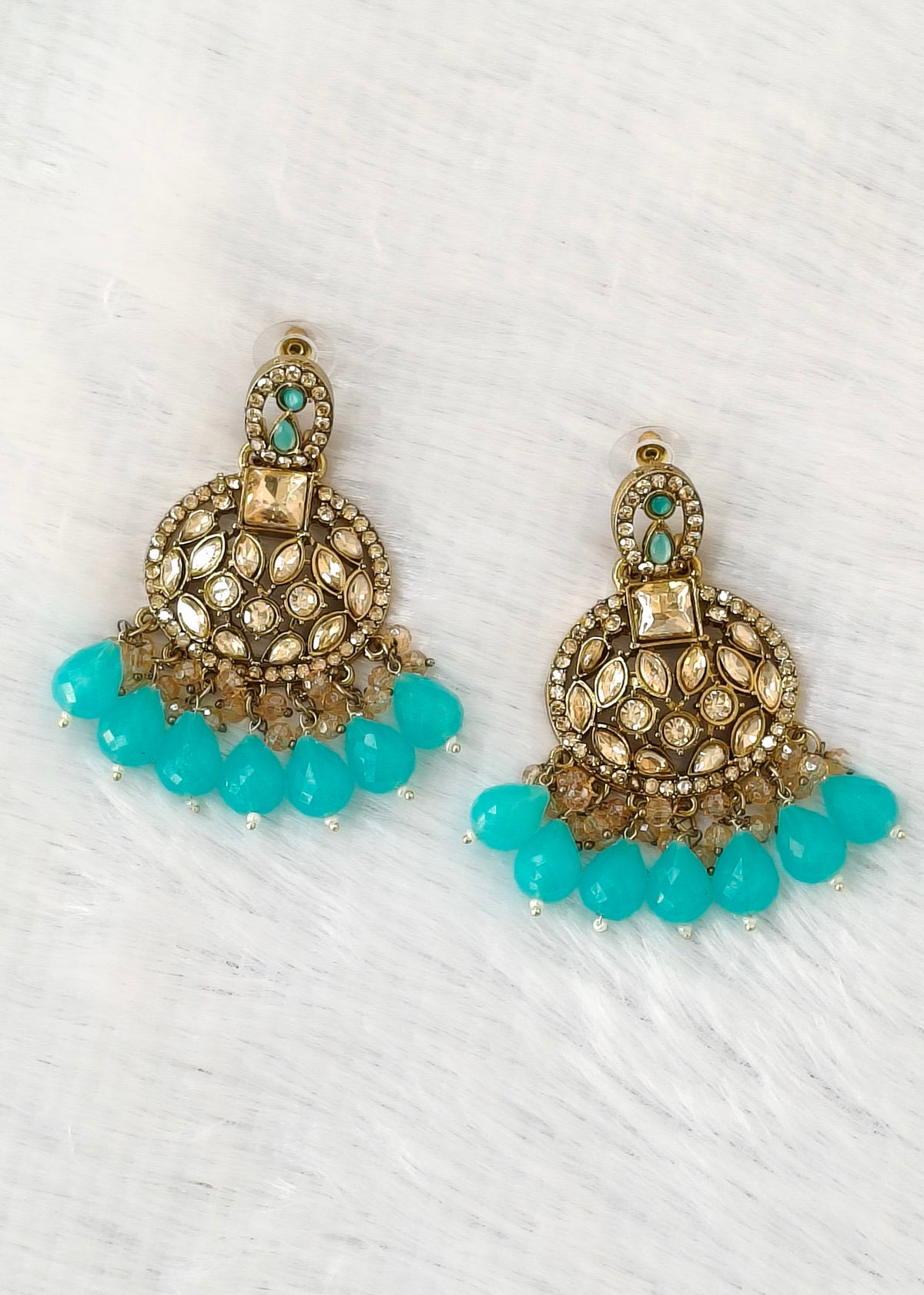 A pair of ornate gold-toned jhumka earrings with turquoise blue bead drops and small clear stones, displayed on a white textured background.