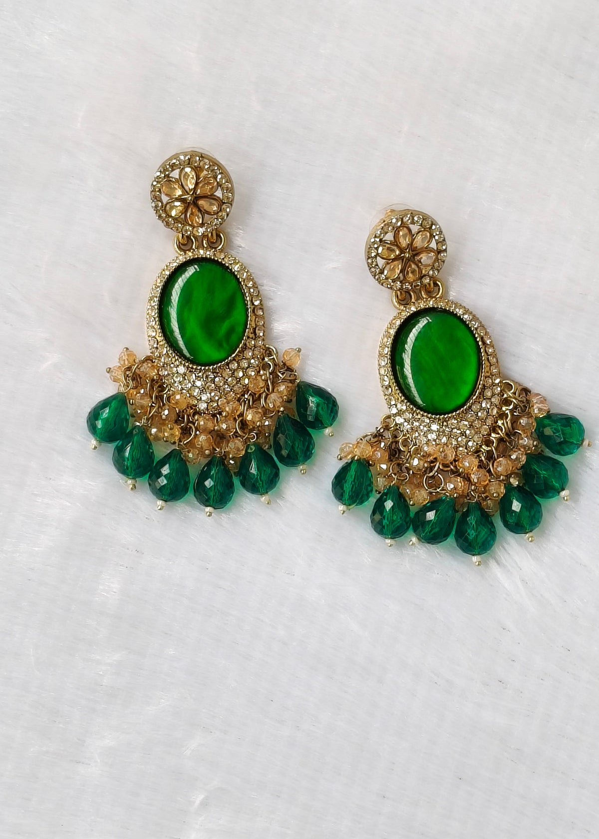 A pair of ornate gold-toned dangle earrings with large oval green stones and dangling green teardrop beads, displayed on a textured white fabric background.