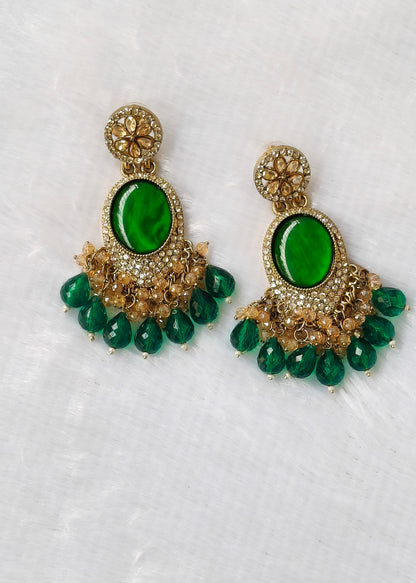 A pair of ornate gold-toned dangle earrings with large oval green stones and dangling green teardrop beads, displayed on a textured white fabric background.