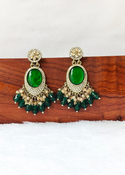 A pair of ornate gold-toned statement earrings with large oval green stones and dangling green bead clusters, displayed on a wooden block against a white background.
