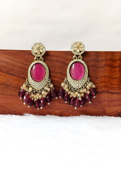 A pair of ornate gold-toned chandelier earrings with oval magenta-pink stone centers and dangling teardrop beads, displayed on a wooden block against a white background.