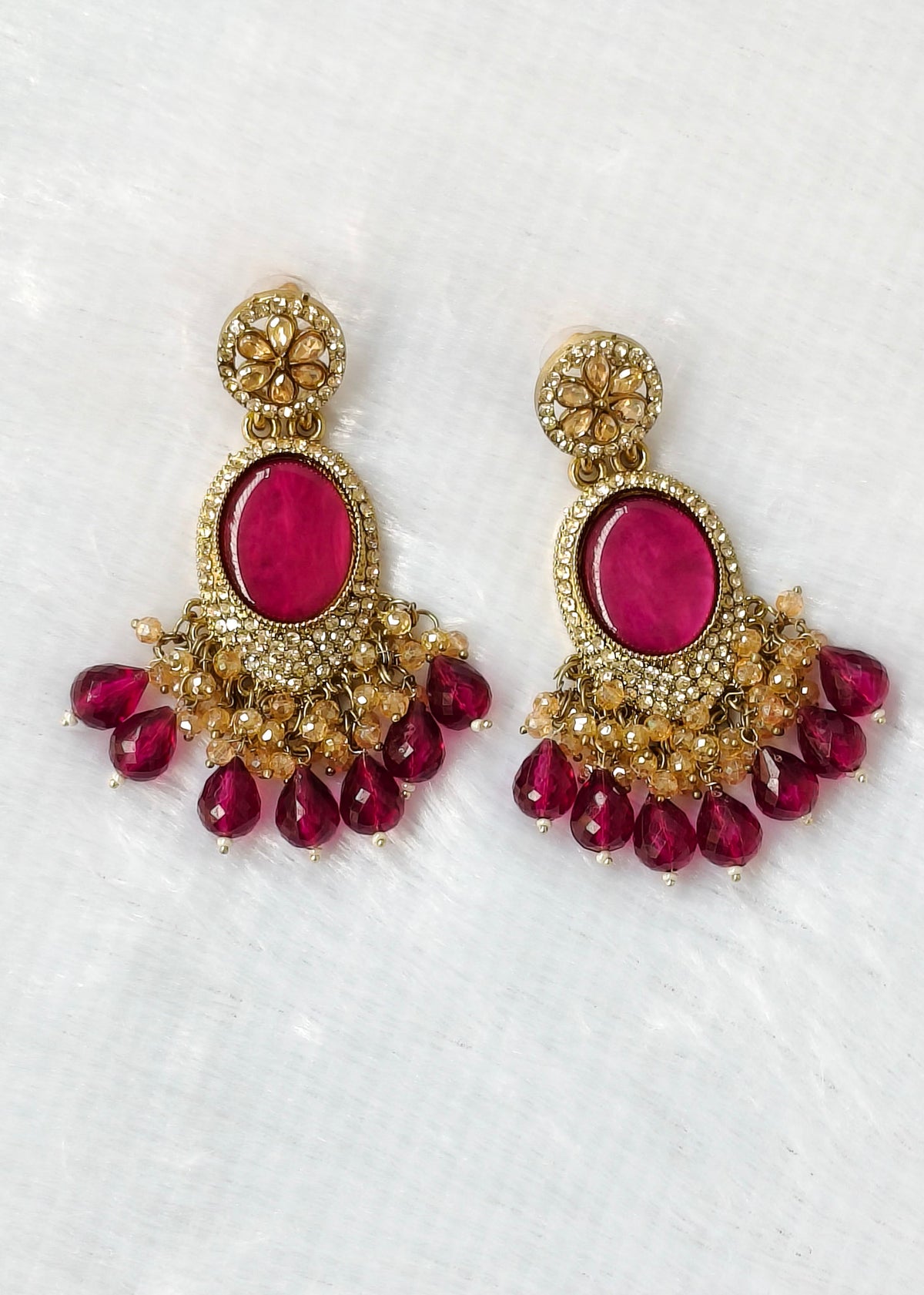 A pair of ornate gold-toned chandelier earrings with oval pink gemstones and dangling teardrop pink beads, displayed on a textured white fabric background.