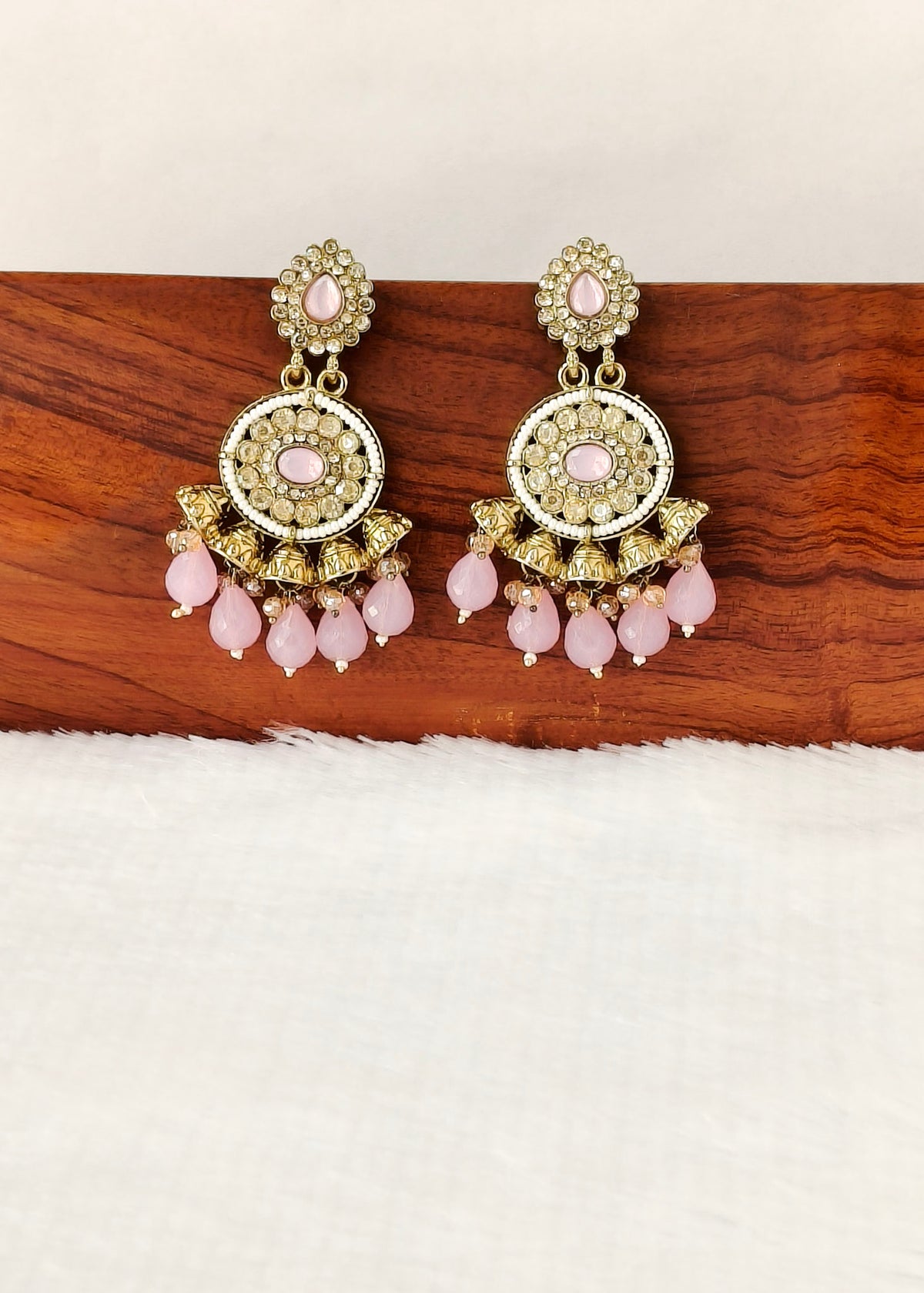 A pair of ornate gold-toned dangle earrings with pink stones and dangling pink bead drops, displayed on a wooden block against a white textured background.