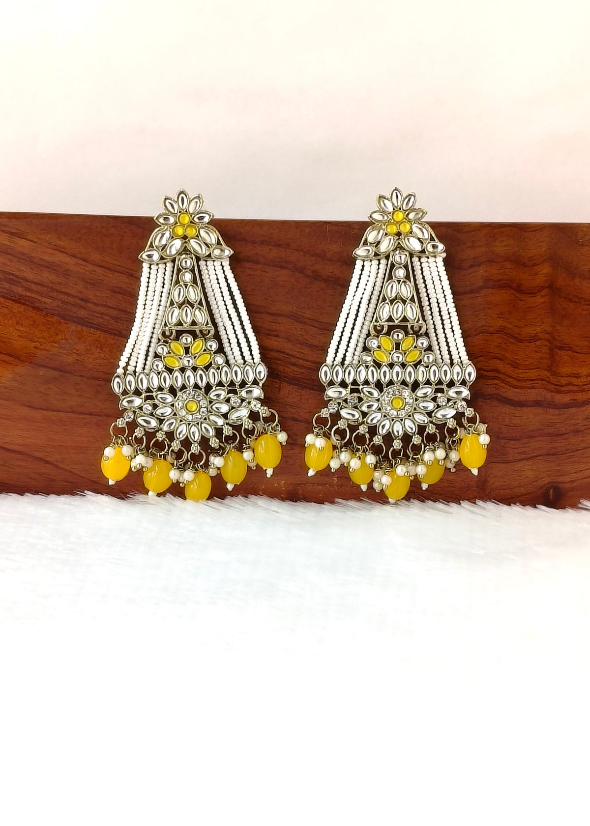 A pair of traditional Indian silver-toned chandelier earrings with yellow bead accents, displayed on a wooden block over a white textured surface.