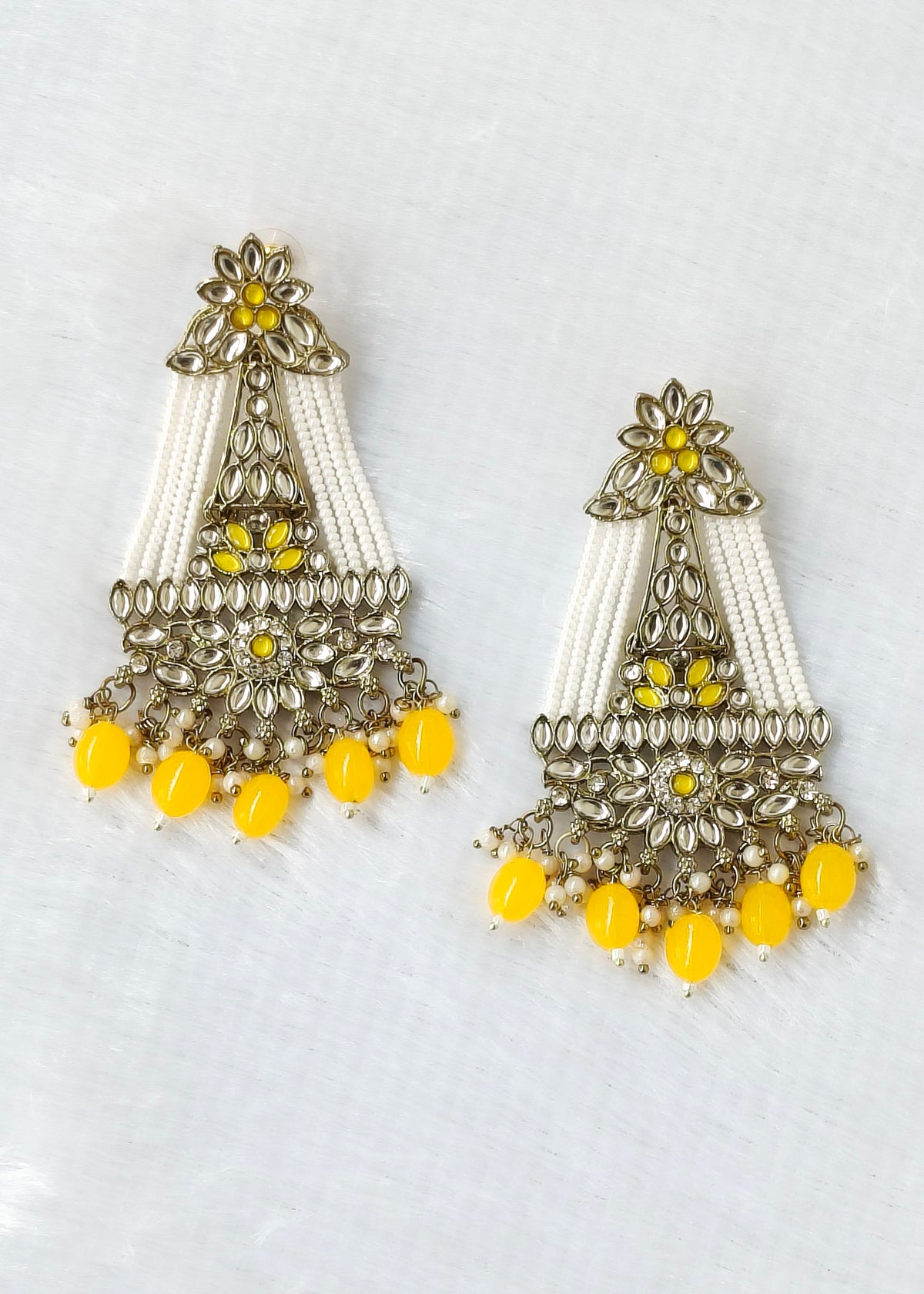 A pair of ornate Indian-style dangle earrings featuring silver-toned filigree, white beaded fringe, and bright yellow bead accents, displayed on a textured white background.