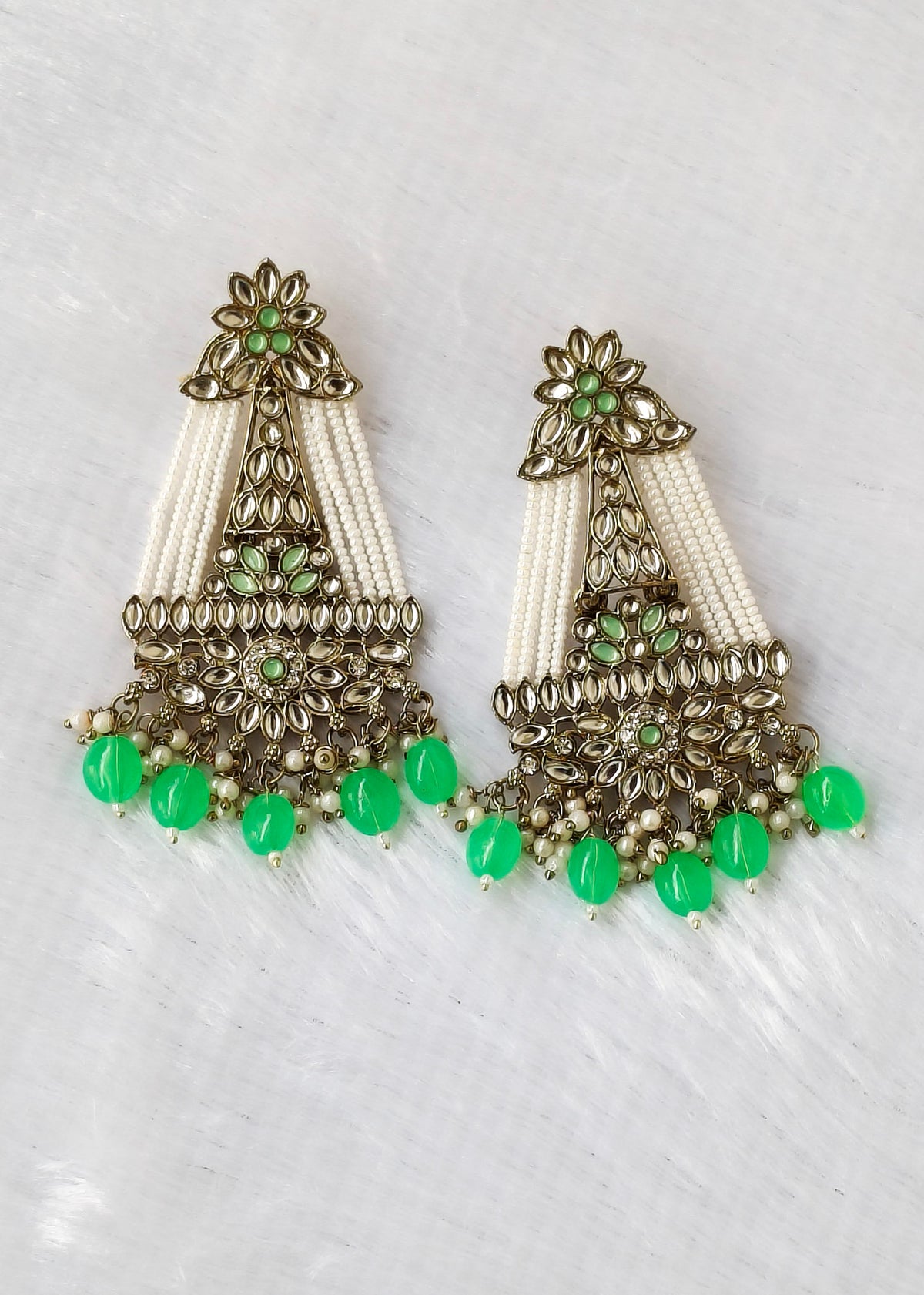 A pair of ornate Indian chandelier earrings with antique bronze metalwork, white seed beads, and mint green gemstone accents, displayed on a white textured background.