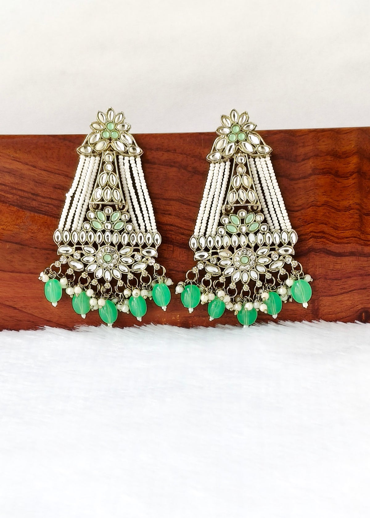 A pair of ornate silver-toned Indian jhumka earrings decorated with clear stones, green bead details, and dangling mint green teardrop beads, displayed on a wooden block over a white surface.