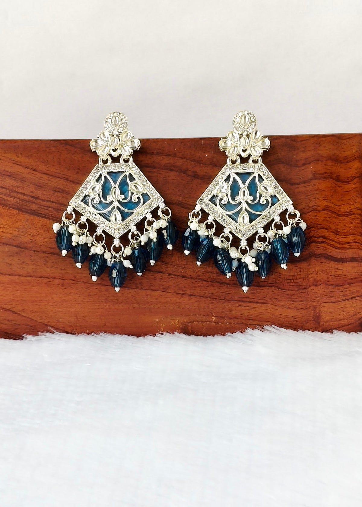 A pair of ornate silver-toned Indian jhumka earrings with blue enamel detailing and dangling dark blue beads, displayed on a wooden block against a white background.