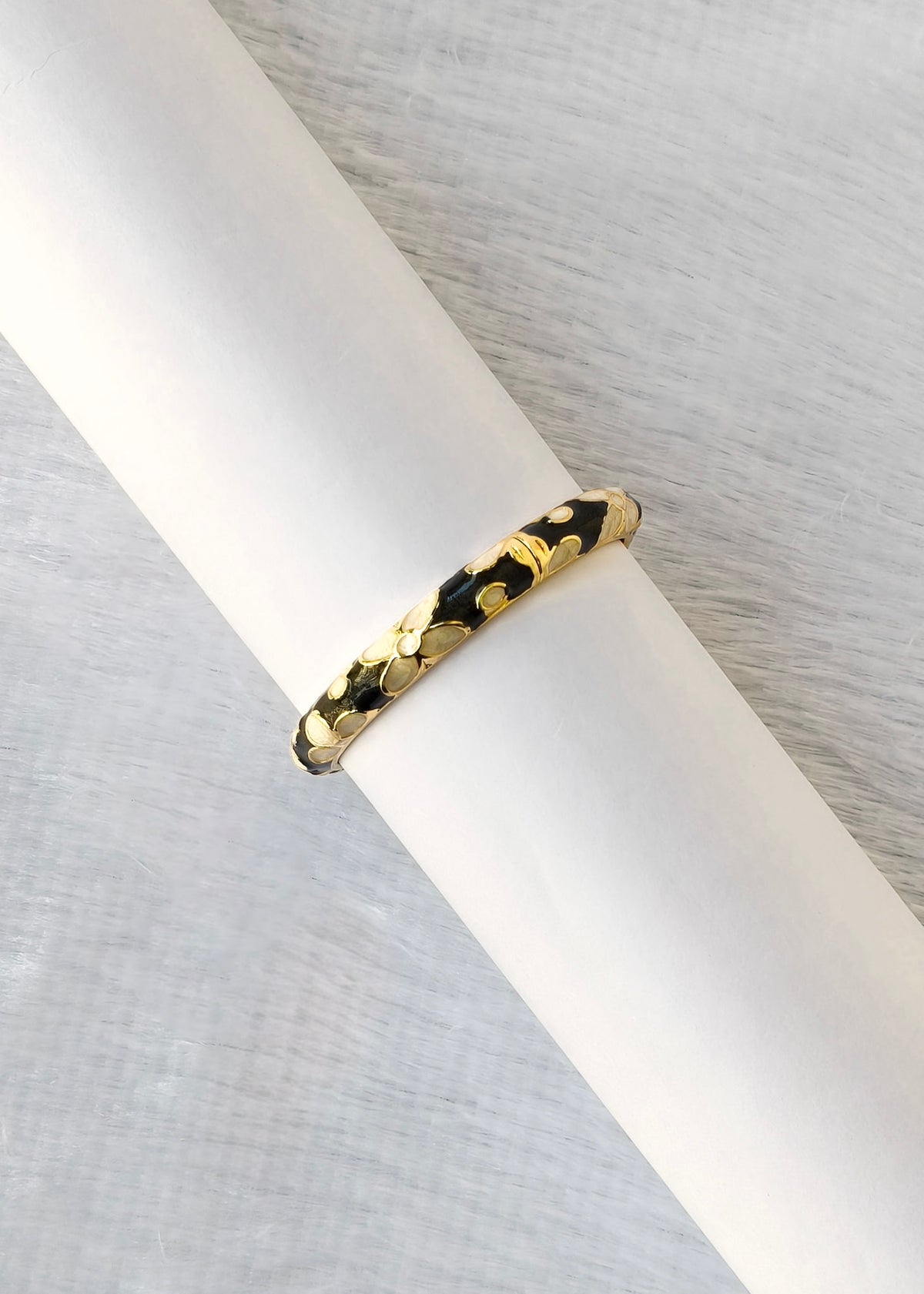 A gold bangle bracelet with a textured, organic leaf-like pattern and black inlay, displayed on a white cylindrical stand against a light gray textured background.