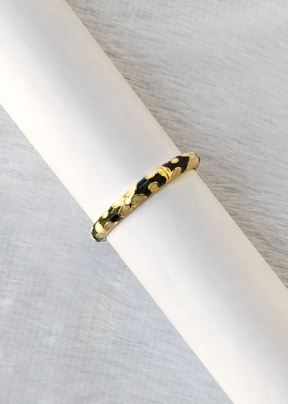 A gold bangle bracelet with a textured, organic leaf-like pattern and black inlay, displayed on a white cylindrical stand against a light gray textured background.