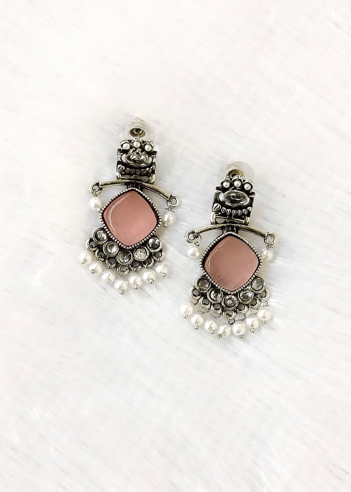 A pair of ornate silver-toned earrings with dusty pink rectangular stone centers and dangling white pearl-like beads, displayed on a white textured surface.