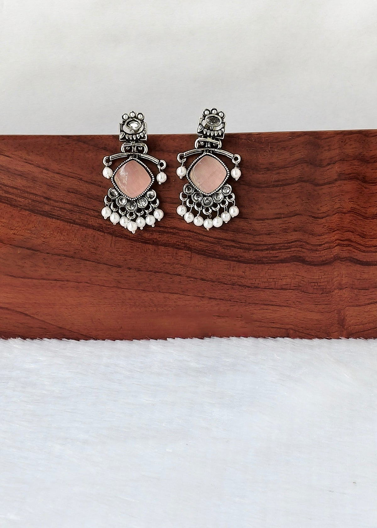 A pair of silver-toned traditional Indian earrings with pink stones and white bead fringe, displayed on a wooden block over a white textured surface.