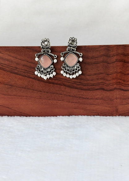 A pair of silver-toned traditional Indian earrings with pink stones and white bead fringe, displayed on a wooden block over a white textured surface.