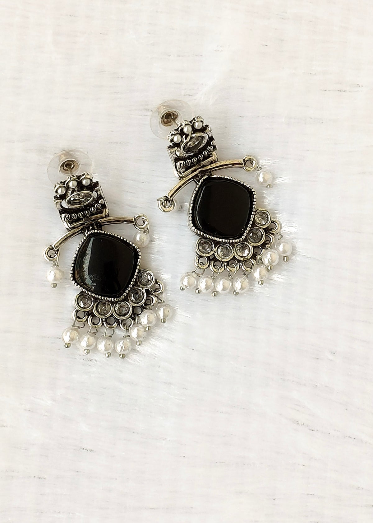 A pair of silver-toned traditional Indian chandelier earrings with black stone centers and small white pearl bead drops, displayed on a light textured white background.