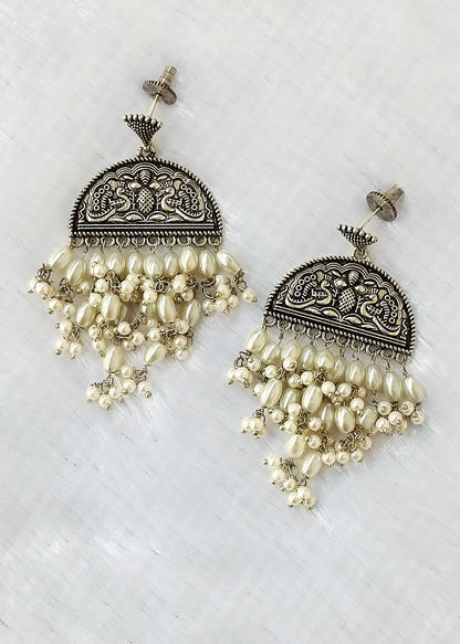 A pair of ornate silver-toned dangle earrings with intricate filigree detailing and cascading cream-colored pearl-like beads, displayed on a textured white surface.