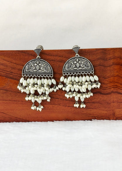 A pair of silver-toned traditional Indian jhumka earrings with intricate filigree work and dangling white pearl-like beads, displayed on a wooden block against a white background.