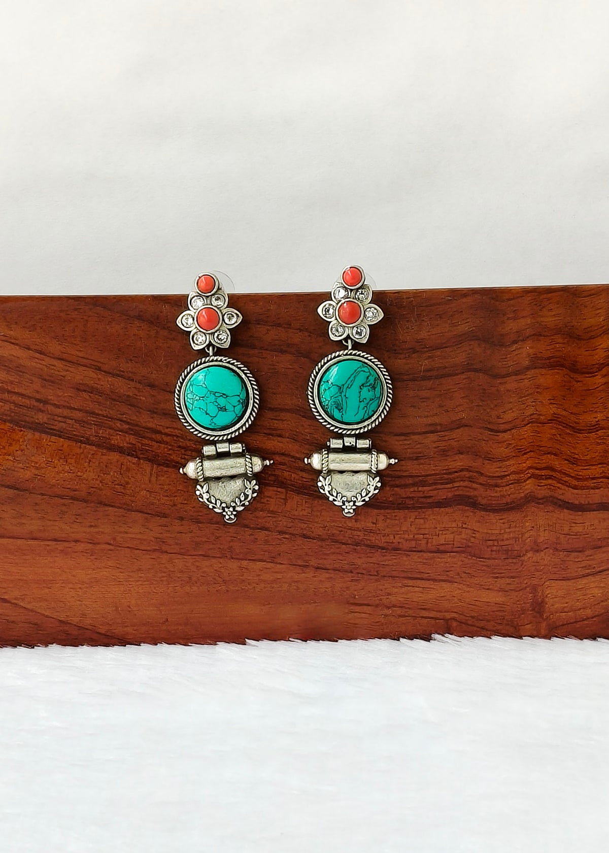 A pair of silver-toned dangle earrings with oval turquoise stones and small coral-red accents, displayed on a wooden block against a white background.