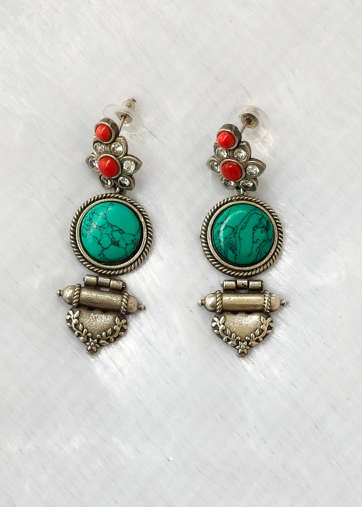 A pair of silver-toned dangle earrings featuring oval turquoise stones, small red coral-like beads, and decorative metalwork, displayed on a white textured background.