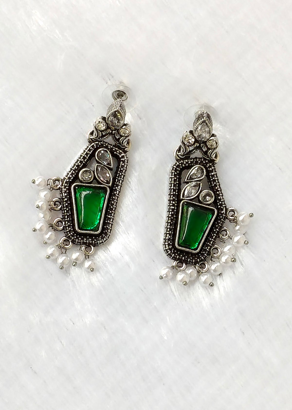 A pair of silver-toned traditional Indian earrings featuring a central green stone and dangling white pearl beads, displayed on a textured white surface.