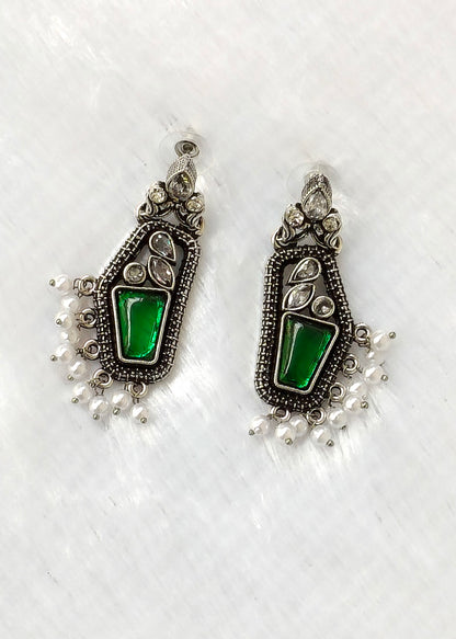 A pair of silver-toned traditional Indian earrings featuring a central green stone and dangling white pearl beads, displayed on a textured white surface.