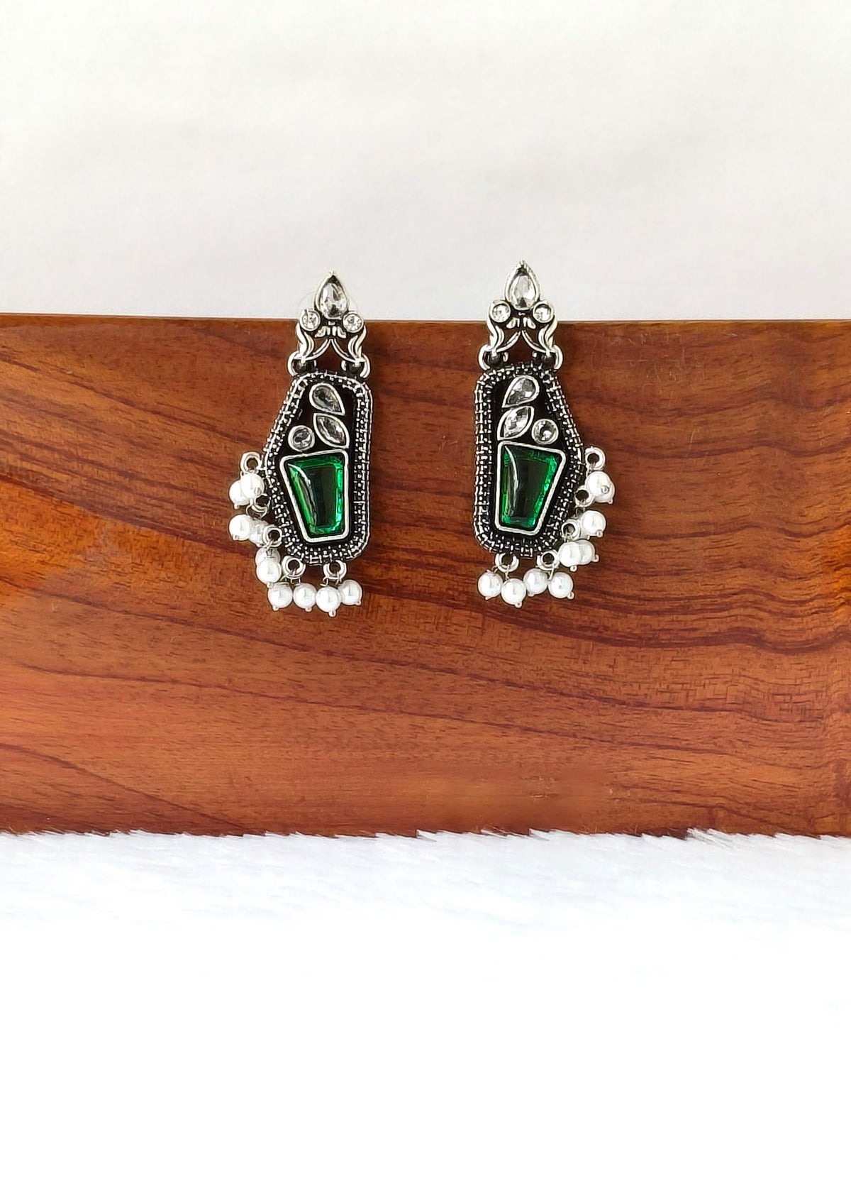 A pair of silver-toned traditional Indian jhumka earrings with green stone inlays and dangling pearl-like beads, displayed on a wooden block against a white background.